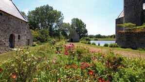 En historisk bygning i sten med stråtækt tag, omgivet af frodigt grønt og mange blomstrende planter.