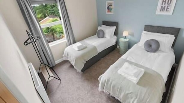 A bedroom with two single beds, a window with curtains, a bedside table with a lamp, and a 'HOME' sign on the wall. - NEC Birmingham Airport Modern Village Retreat, Sleeps 7, Free Parking (Hampton in Arden)