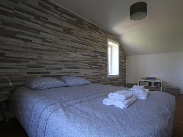 Un dormitorio con techo inclinado, una cama con colcha gris, dos almohadas y una ventana. Hay una mesita de noche blanca pequeña con una lámpara y una pila de toallas dobladas.
