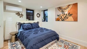 A bedroom with a bed, a bedside table, a wall-mounted air conditioner, a window, and a decorative wall piece. - Lioness Downtown Den (Denver)