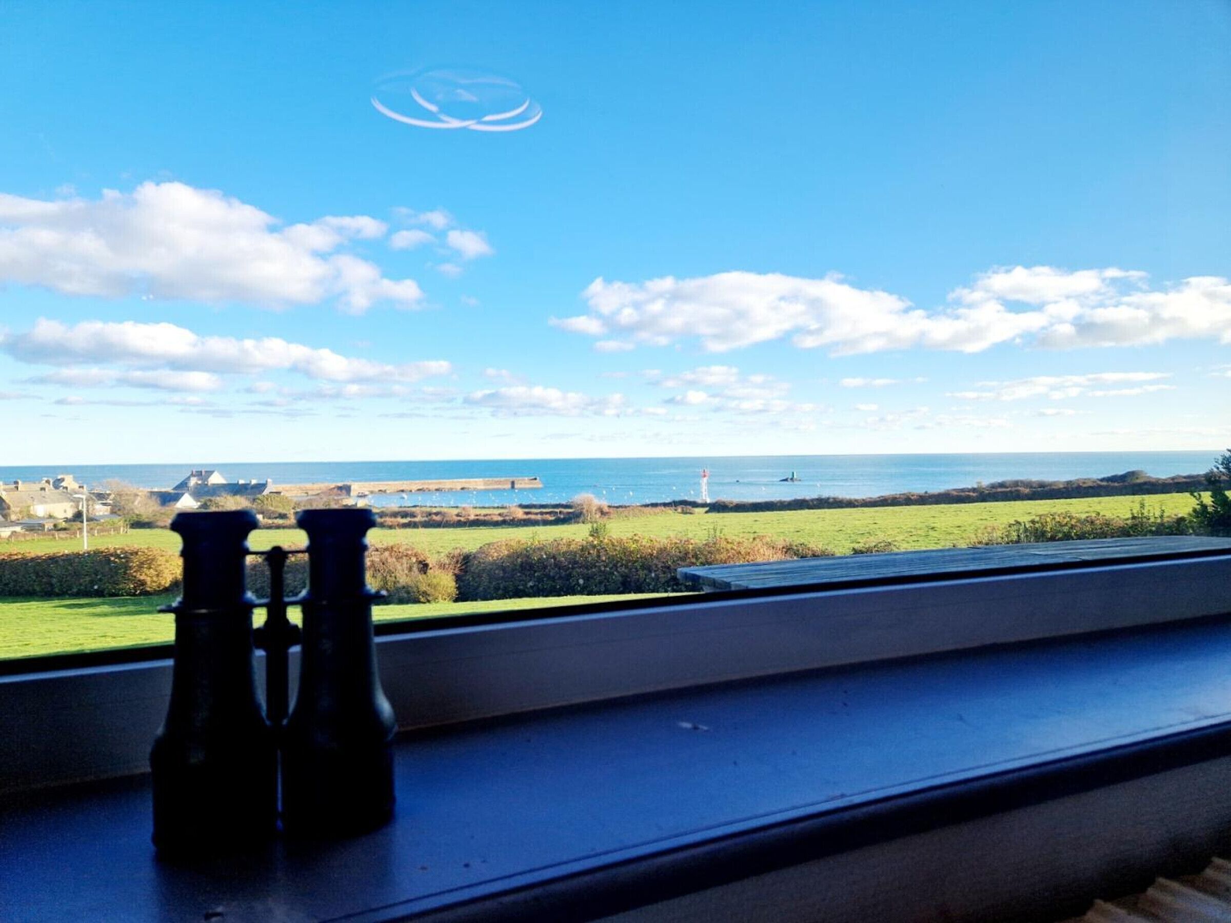 Une vue par la fenêtre sur un paysage côtier, avec un phare, des champs verdoyants et un ciel dégagé.