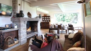 A cozy living room with a stone fireplace, a television mounted on the wall, and a dining area with a large window. - Spacious house with sea view, fireplace and garden (La Hague)