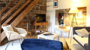 Ein gemütliches Wohnzimmer mit einer Steinwand, einer Holztreppe, einem Fernseher, Korbsesseln, einem blauen Sofa und einer Stehlampe.