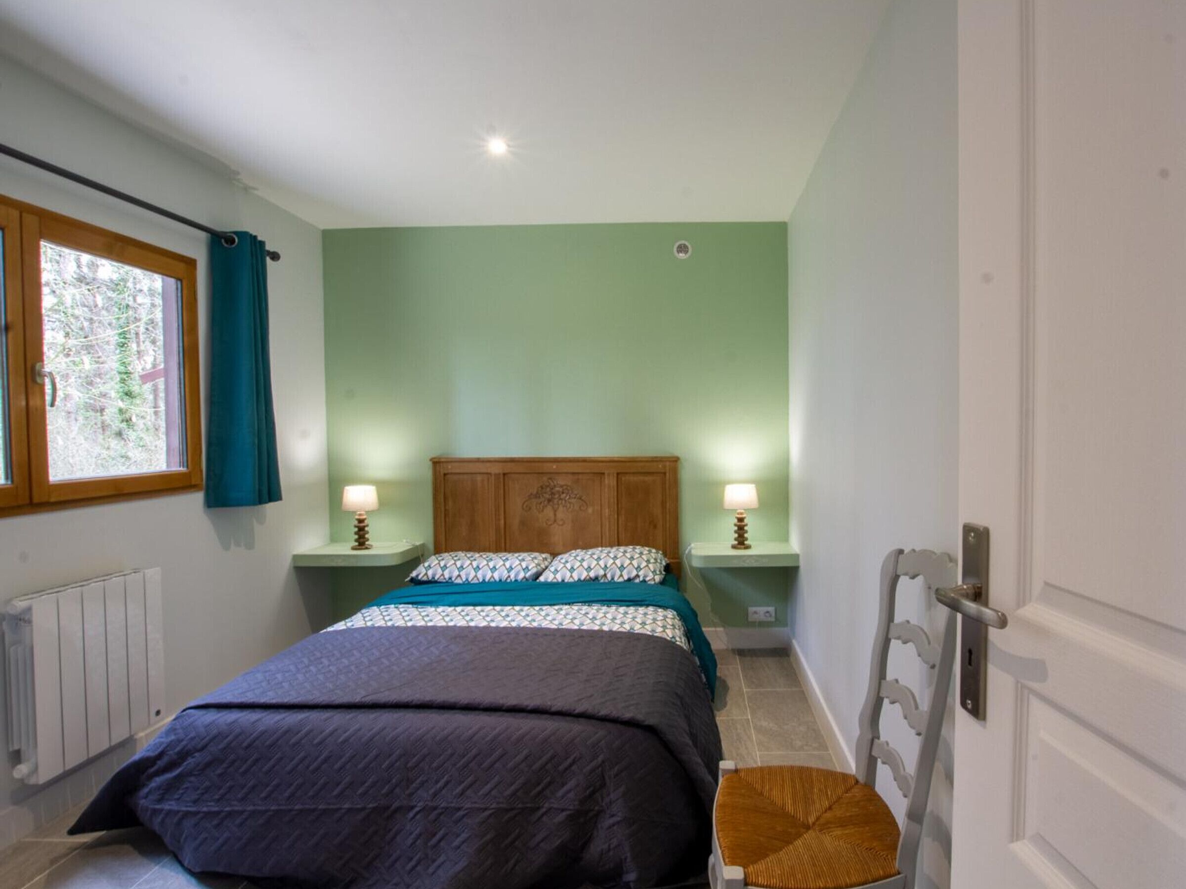 A bedroom with a bed, two bedside tables with lamps, a window with curtains, and a radiator.