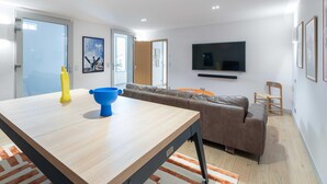 A modern living room with a wooden dining table, a sofa, and a television mounted on the wall. - Blueberry of the Valley - House With Swimming Pool (Saint-Pierre-Azif)