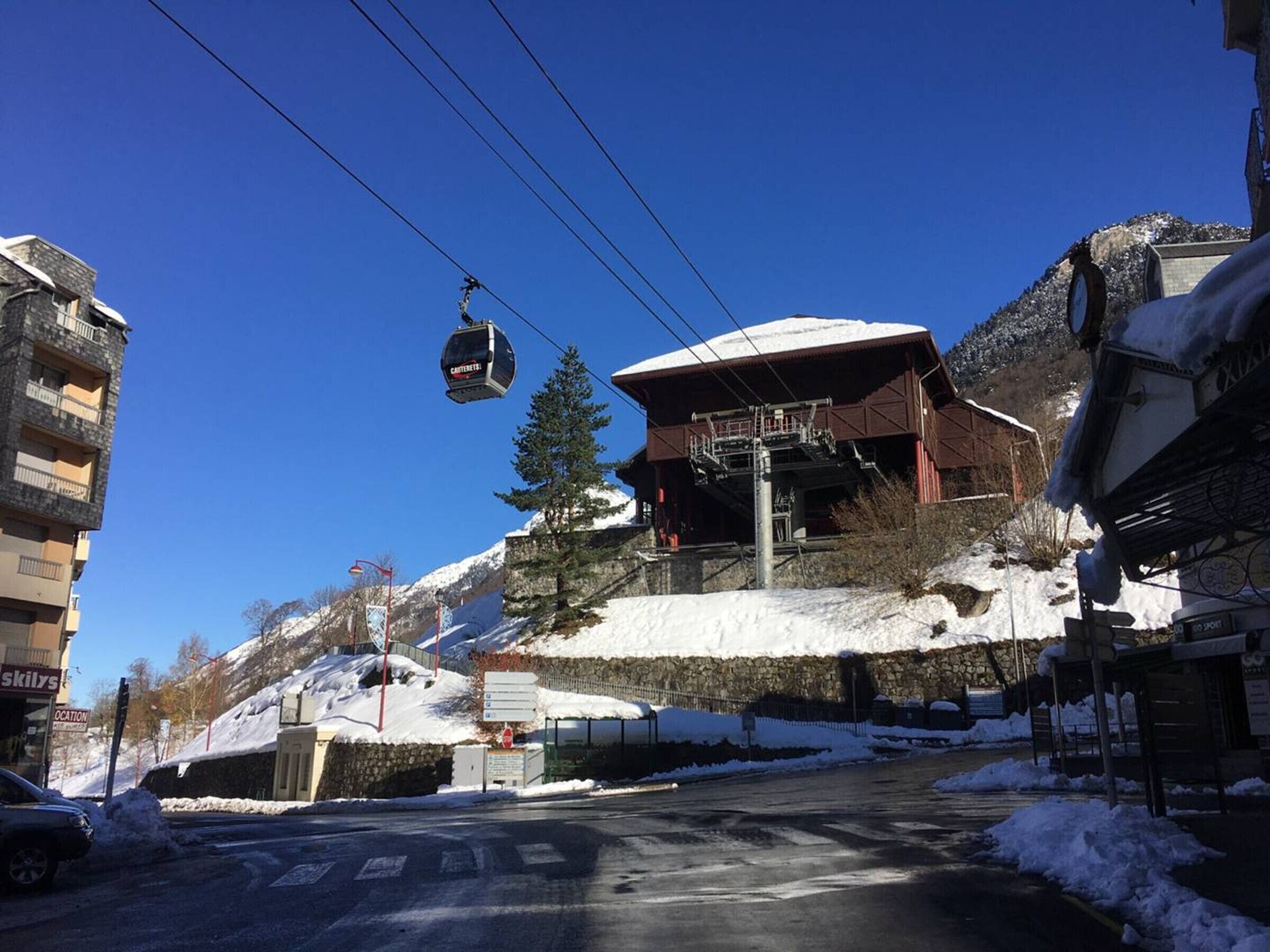 A ski lift chair suspended above a building with a snow-covered roof, a stone wall, and a sign indicating a ski location.