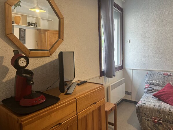 A bedroom with a wooden dresser, a desk with a computer, a red clock, a window with curtains, and a bed with a patterned duvet.