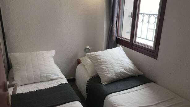 A small, compact room with two single beds, each with white bedding and a dark headboard. There is a window with a view of a balcony.