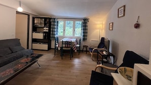 A cozy living room with a sofa, a dining table with chairs, a TV, and a kitchen area with cabinets and a window. - Cauterets: Central T2 Apartment, 4-5 pers (Cauterets)