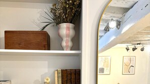 A room with a white wall, a wooden chest, a vase with dried flowers, a small vase with a yellow flower, and a mirror reflecting a dining area with a table, chairs, and a sofa. - Apartment Saint Germain des Pres Bon Marche (Paris)