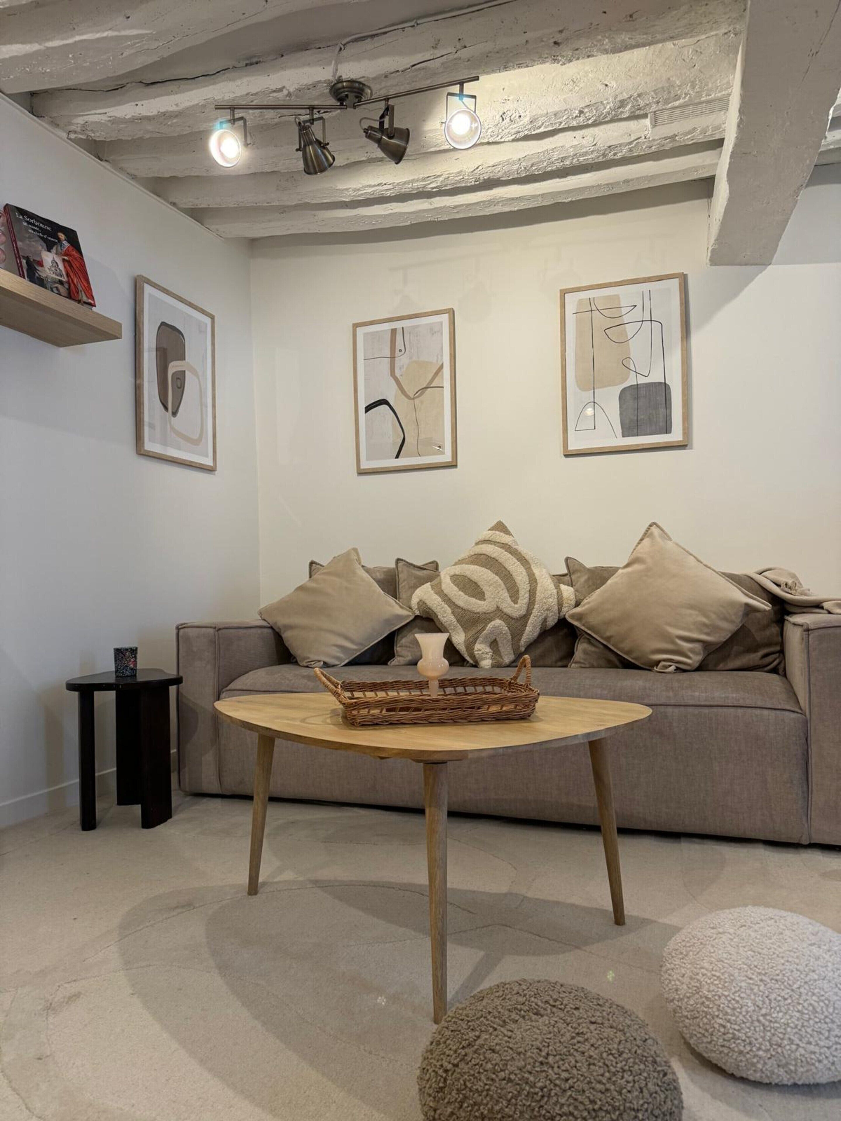 Una sala de estar moderna con un sofá gris, una mesa de centro redonda de madera y una mesita auxiliar negra. Hay pinturas abstractas en las paredes y un candelabro en el techo.