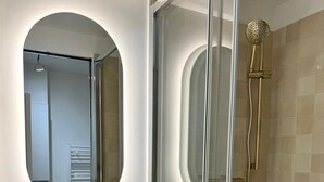 A modern bathroom with a round white sink, a gold faucet, a glass-enclosed shower with a gold showerhead, and a mirror with a circular frame. - Apartment Saint Germain des Pres Bon Marche (Paris)