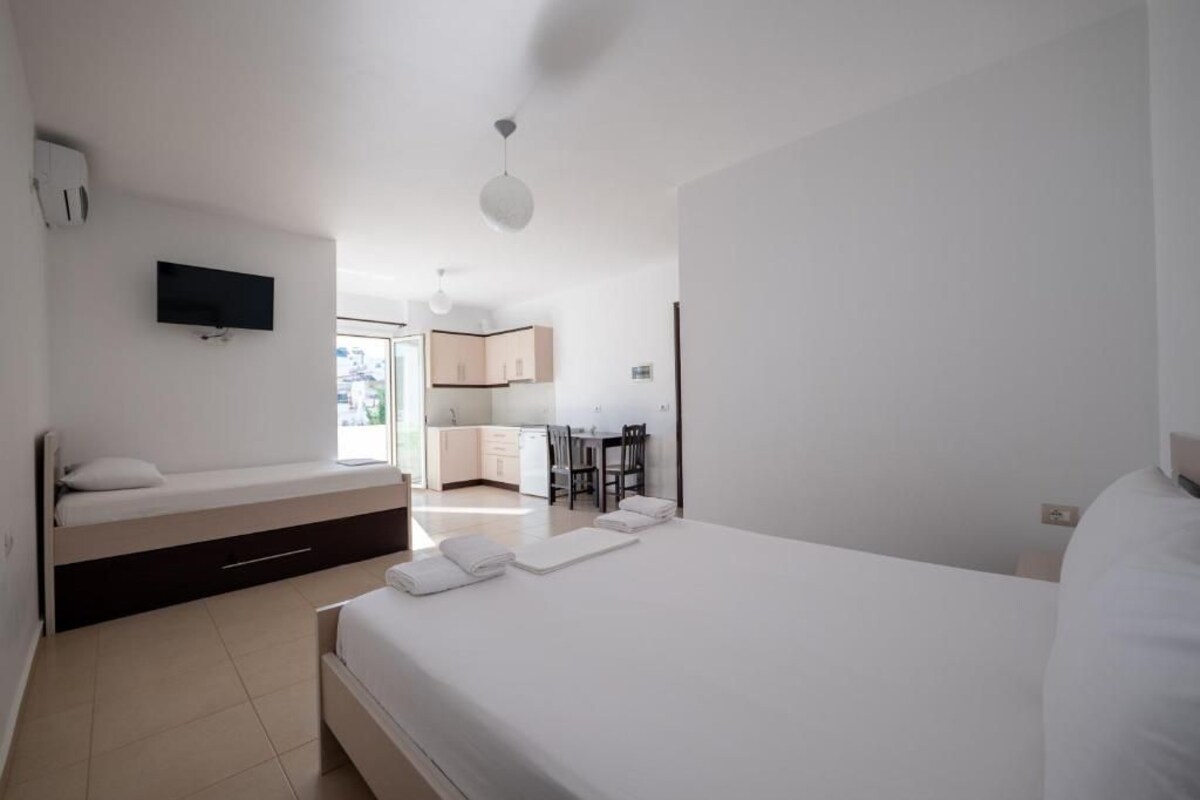 A modern bedroom with a large bed, a built-in bench with storage, a TV mounted on the wall, and a view into the kitchen area.