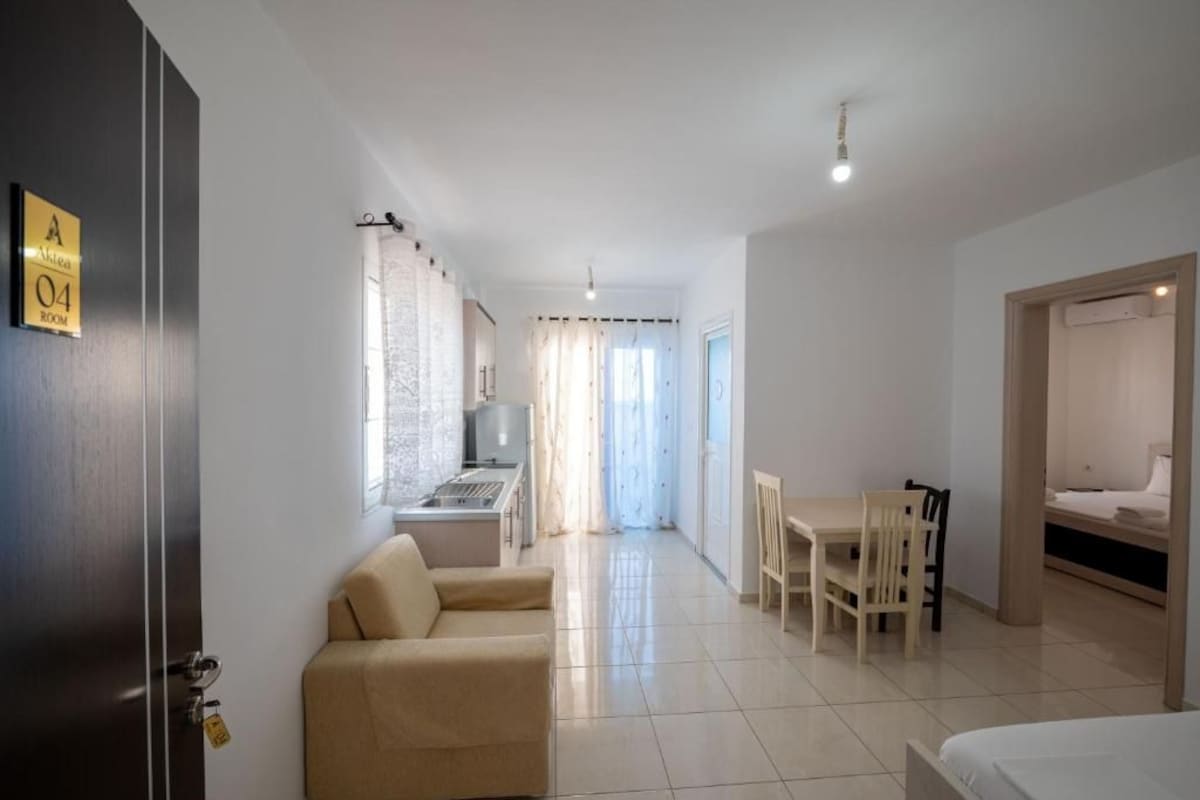 A hotel room with a bed, a sofa, a dining table with chairs, a kitchen area, and a bathroom.