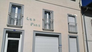 Un immeuble avec une enseigne « Les Pins » au-dessus de l’entrée. Il y a trois fenêtres avec des garde-corps en métal, dont une est fermée par un volet. L’immeuble a une façade beige et une porte bleue.