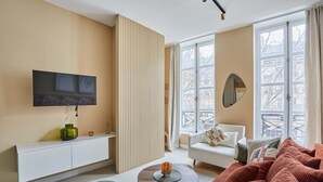 A modern living room with a sofa, a coffee table, a TV mounted on the wall, and a window with a view of buildings. - Sublim Apartement in the Center of Paris/le Marais (Paris)