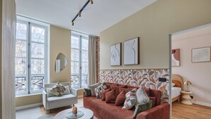 A living room with a sofa, armchair, and a round coffee table. There are large windows with a view of buildings outside. - Sublim Apartement in the Center of Paris/le Marais (Paris)