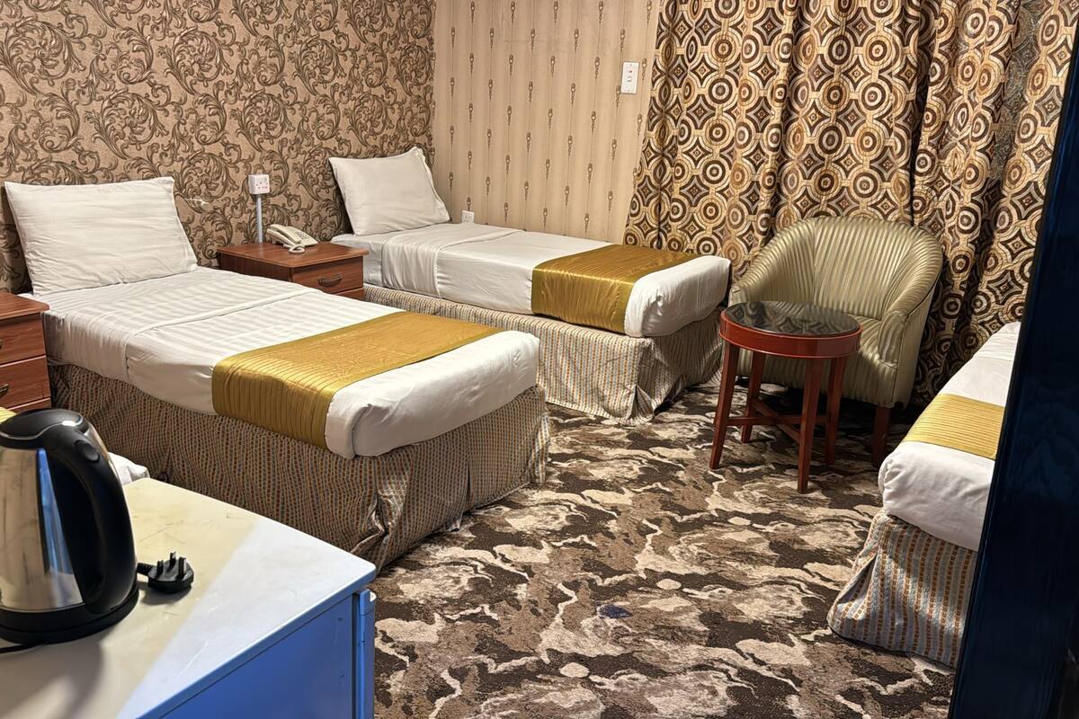 A hotel room with two beds, a chair, a desk with a kettle, and patterned wallpaper.