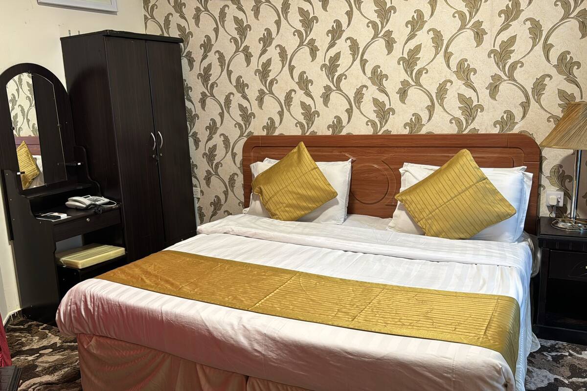A neatly made bed with white and gold bedding, a wooden headboard, and two gold pillows. A black wardrobe with a mirror is to the left, and a nightstand with a lamp is to the right.