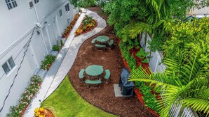 A courtyard with a green lawn, two round tables with chairs, and a white building with windows and a white fence. - Stylish Miami Studio | Parking+Gym | Near Aventura (Miami)
