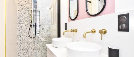 Un baño moderno con ducha de vidrio, dos lavamanos con detalles dorados y un dispensador de jabón negro.
