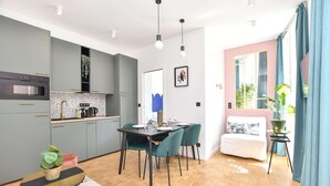 Eine moderne Küche mit grauen Schränken, ein Essbereich mit Tisch und Stühlen, ein Bett mit weißer Daunendecke und ein Fenster mit Vorhängen.
