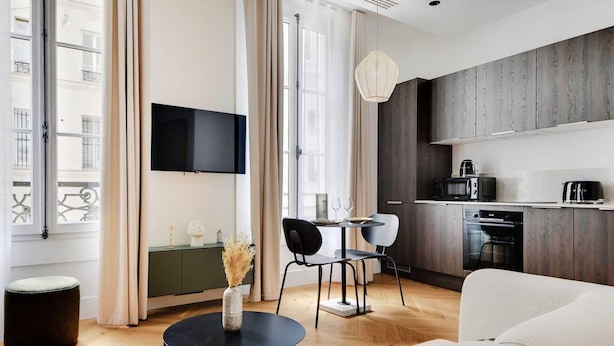 Un soggiorno moderno con un divano, un tavolino rotondo e una TV fissata al muro. C'è un'area pranzo con tavolo e sedie, e un angolo cottura dotato di forno a microonde e macchina del caffè.