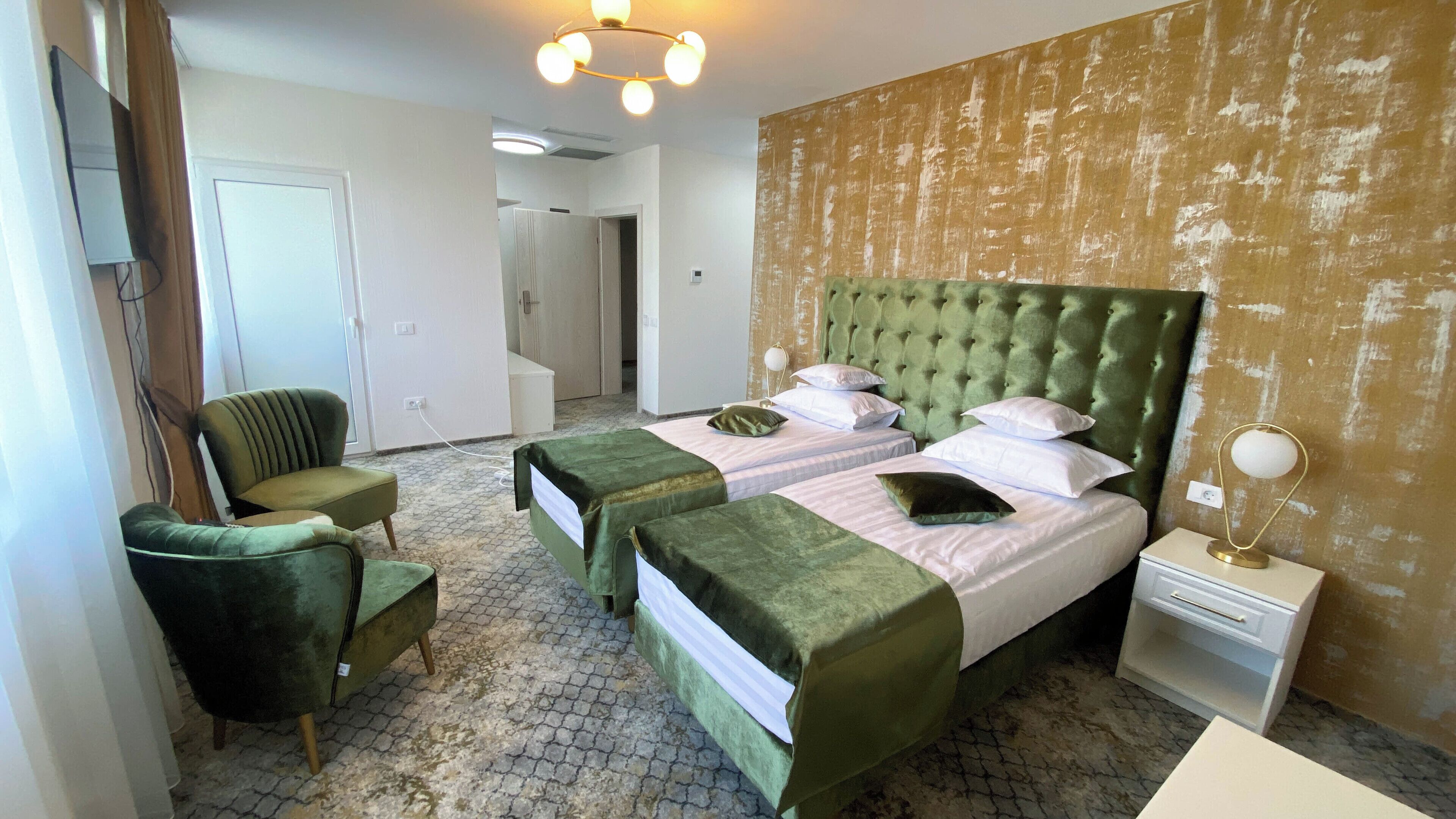 A hotel room with two beds, a green armchair, a bedside table with a lamp, and a door leading to another room.