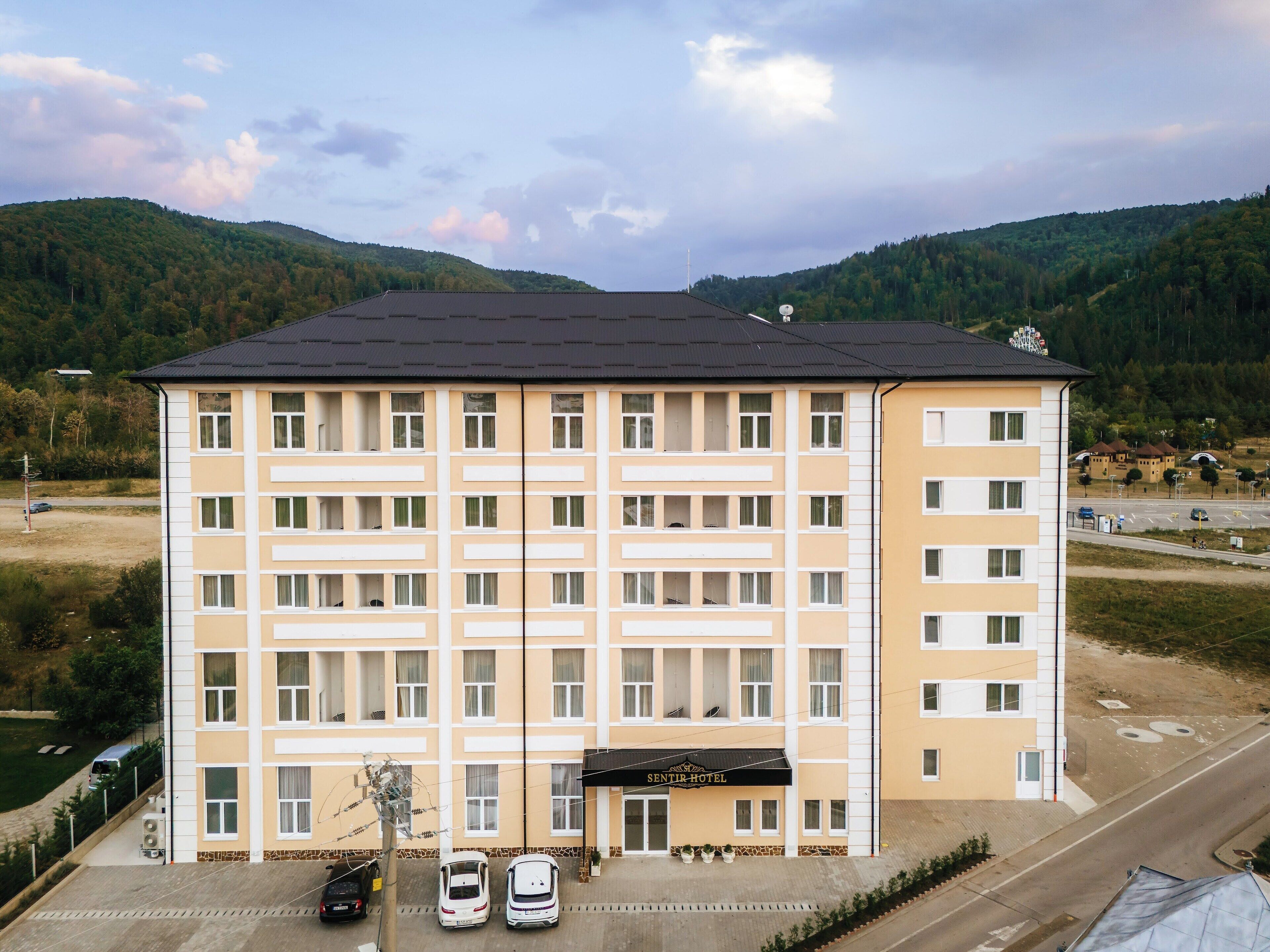 A multi-story hotel with a flat roof, a central entrance, and a parking area with several cars.