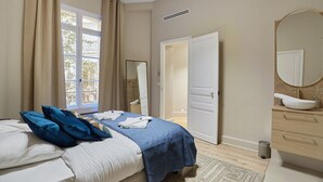 Ein Schlafzimmer mit Bett, einem Fenster mit Vorhängen, einem Badezimmer mit Waschbecken und Spiegel sowie einer Tür zu einem anderen Raum.