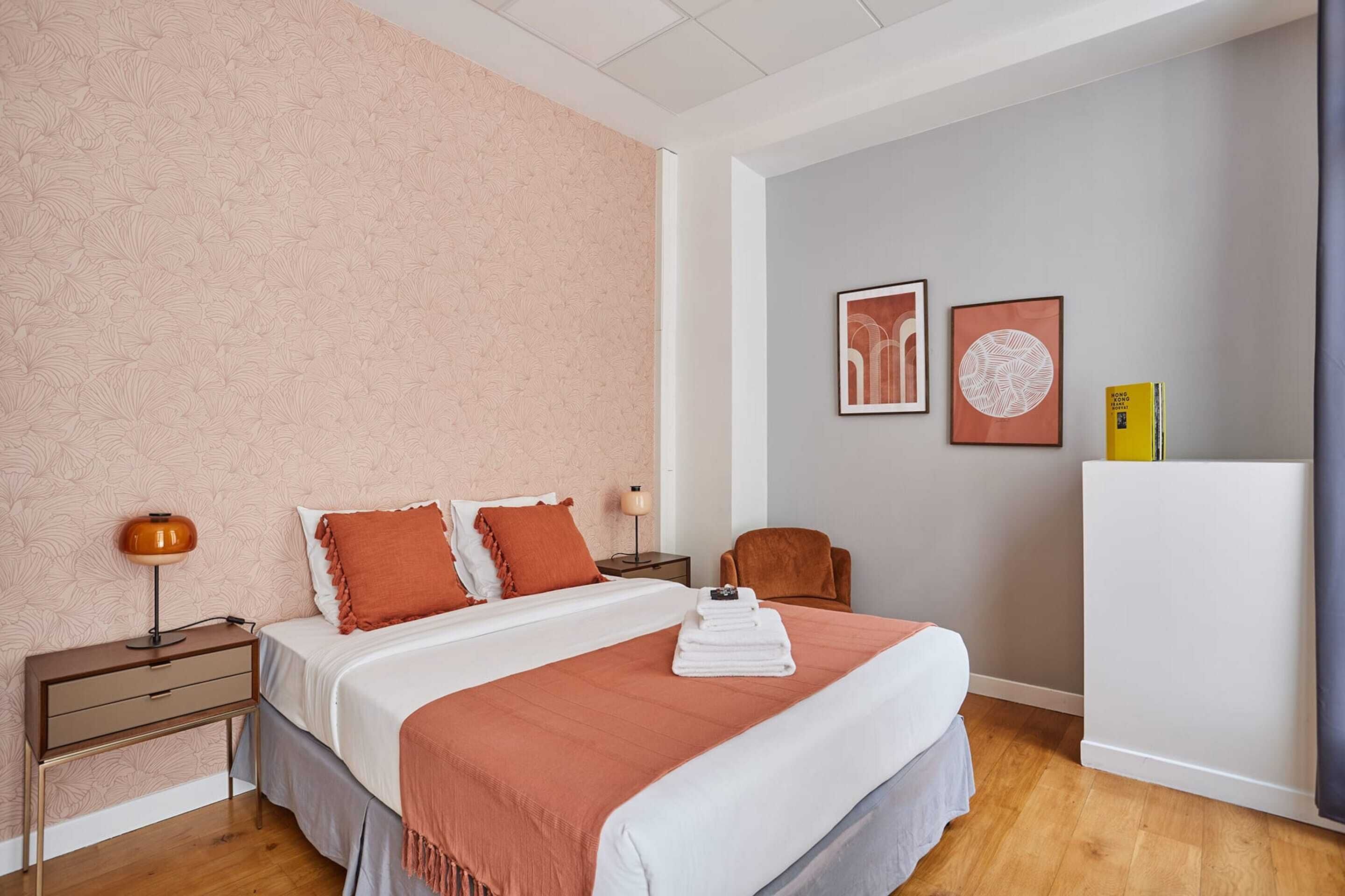 A neatly arranged hotel room with a bed, bedside table, lamp, chair, and artwork on the wall.