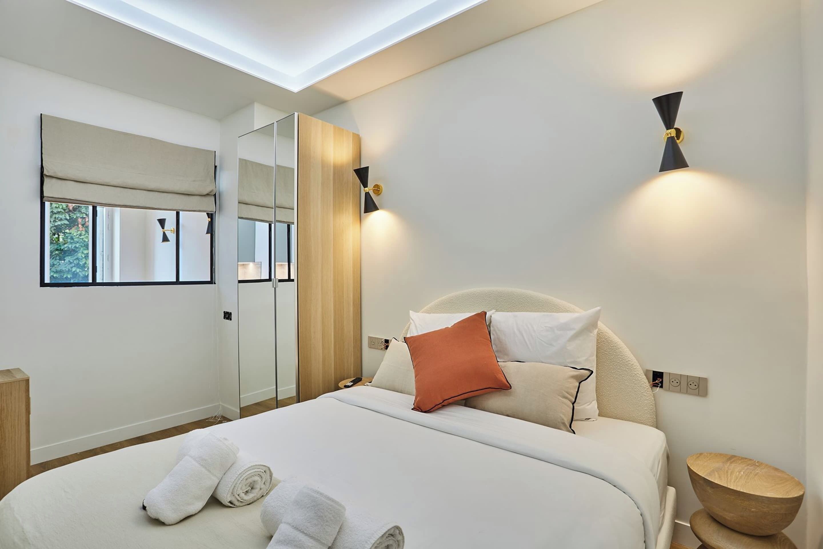 A modern hotel room with a large bed, a mirror, a bedside table, and a wall-mounted light fixture.