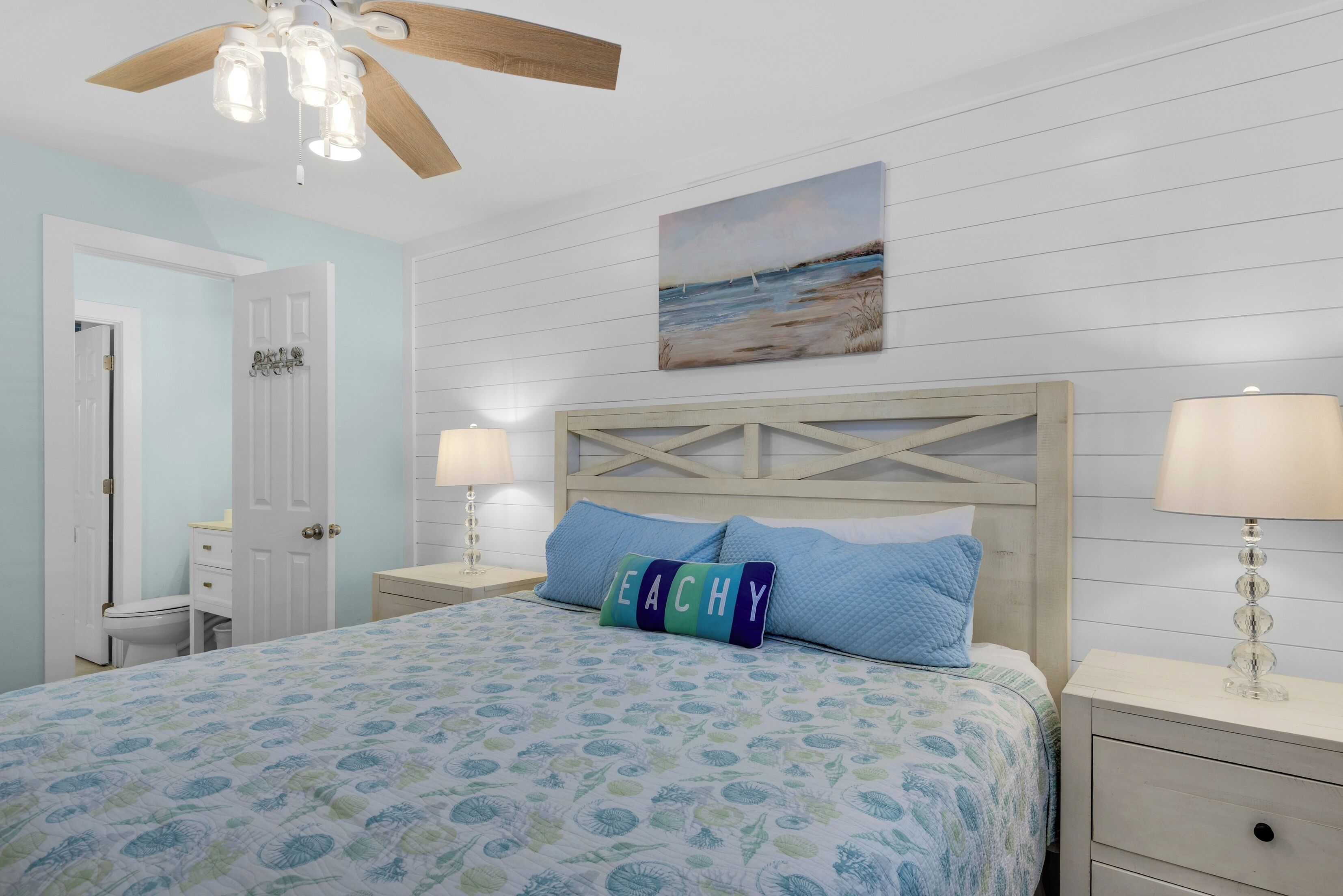A bedroom with a bed, two bedside tables with lamps, a ceiling fan, a door, and a wall-mounted painting.