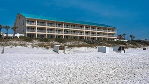 A large, multi-story hotel with a flat roof and balconies, located on a sandy beach with palm trees in the background.
