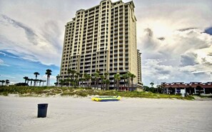 A tall hotel with multiple balconies, a sandy beach in the foreground, and palm trees.