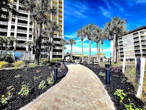 A pathway leading to a building with multiple balconies, surrounded by palm trees and a clear sky.