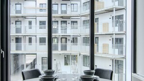 Un espace repas comprenant une table et des chaises, de grandes fenêtres et une vue sur un immeuble à plusieurs étages.
