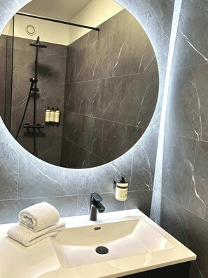 A bathroom with a round mirror, a white sink, a towel, and a shower with a handheld showerhead. - B2 - One Bedroom Apartment with Balcony (Reykjavík)