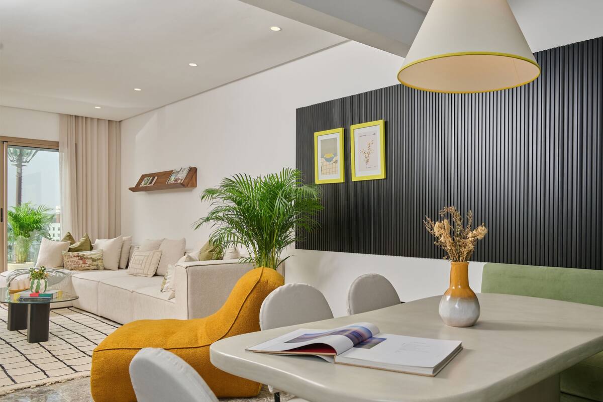 A modern living room with a sofa, a dining table with a book, a potted plant, and a wall with vertical panels.