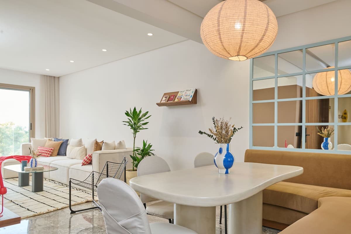A modern living room with a sofa, a dining table, and a coffee table. There is a large window with a view of the outdoors.