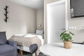 A modern hotel room with a bed, a chair, a dining table, a plant, and a TV mounted on the wall.