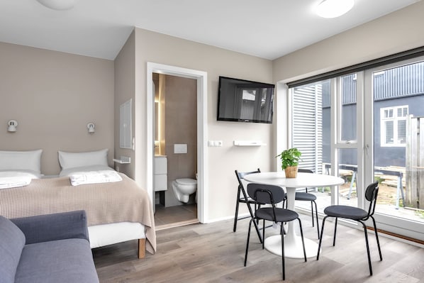 A modern hotel room with a bed, a dining area with chairs, a TV mounted on the wall, and a large window offering a view.