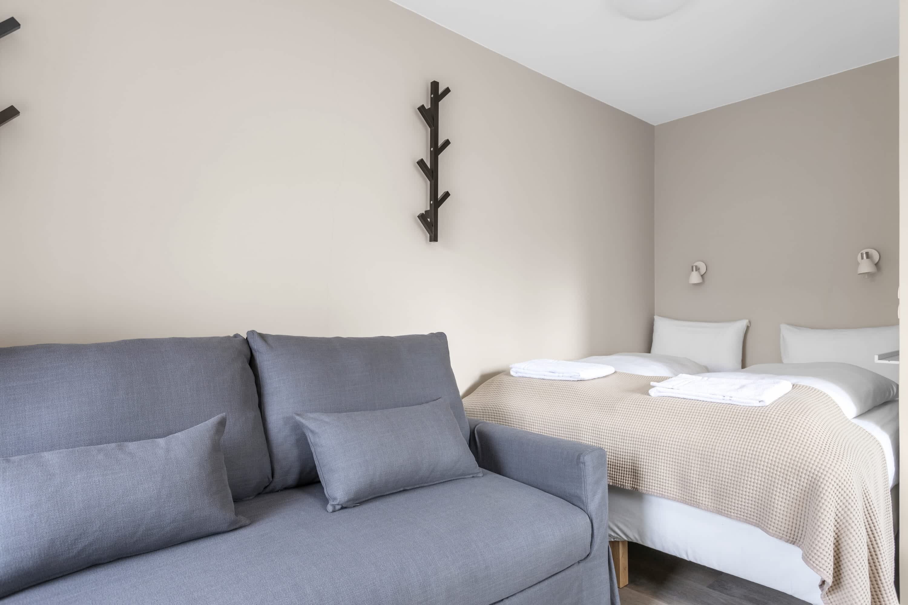 A neutral-colored room with a grey sofa, a bed with white bedding, and a wall-mounted light fixture.
