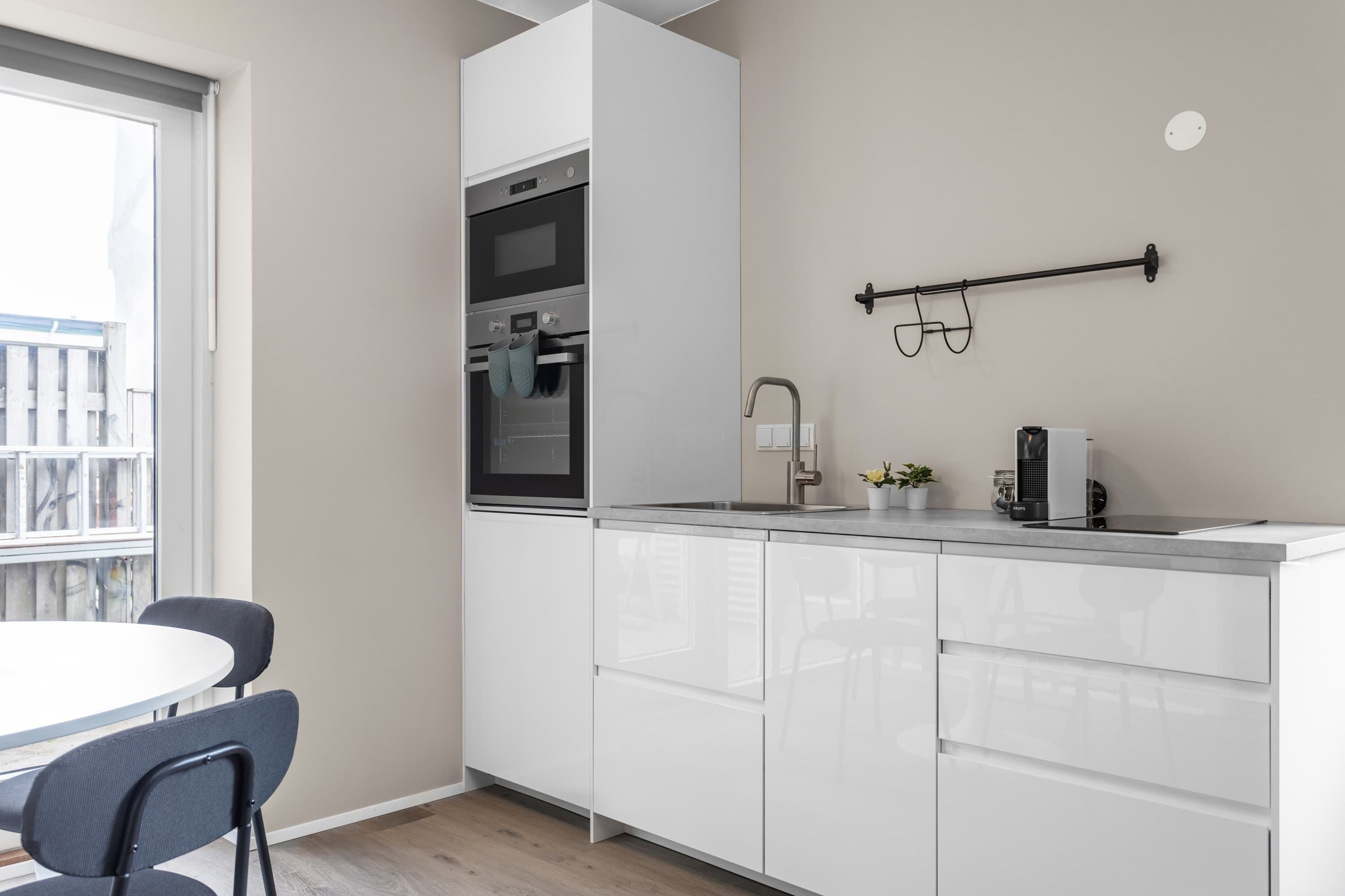 Een moderne keuken met witte kastjes, een inbouwoven en een raam met uitzicht op een balkon.