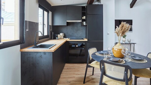 A compact kitchen with dark cabinetry, a wooden countertop, and a dining area with a table set for two. - Apartment AC 2 Bedrooms Le Marais Beaubourg (Paris)