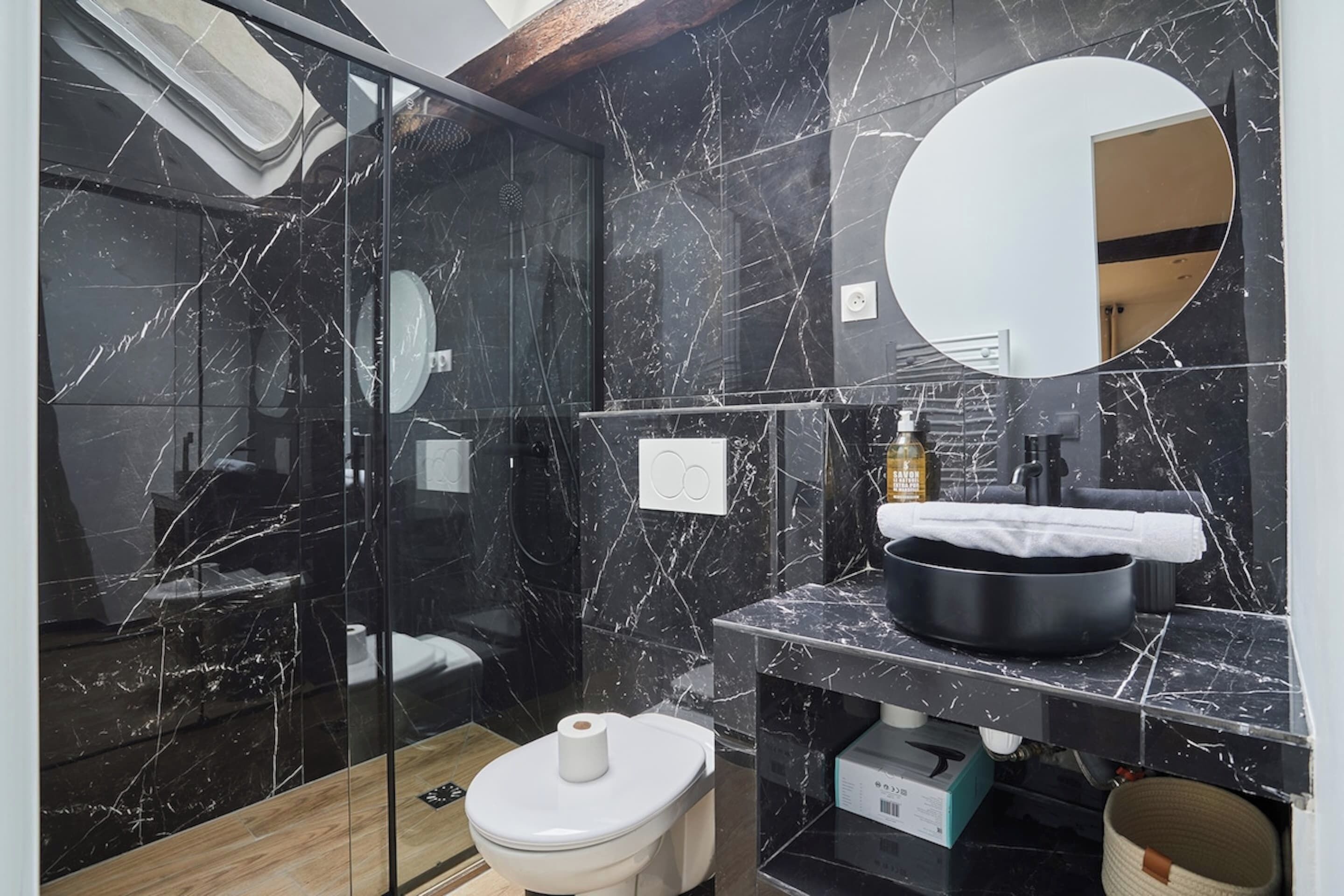 Een moderne badkamer met een douchecabine van glas, een betegelde muur van marmer, een ronde spiegel en een zwart wastafelmeubel met een handdoek.