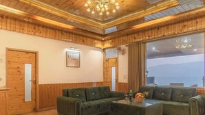 A cozy room with wooden walls and ceiling, a green sofa set, a glass coffee table, and a large window offering a view of the mountains. - The Castle Villa Hill View kasauli (Kasauli)