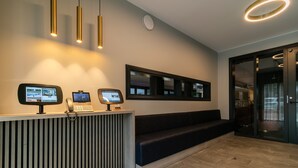 A reception desk with a computer monitor, a telephone, and a digital screen. There is a bench and a glass door leading to another room.