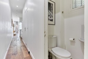 A narrow hallway with a wooden floor, white walls, and a white toilet with a framed picture above it.
