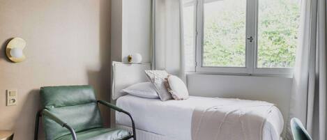 Una cama individual con colchón y almohadas blancos, una silla verde y una ventana con cortinas.